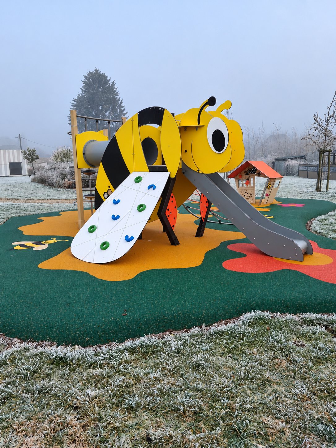 Structure de jeux en forme de grosse abeille jaune et noire avec toboggan et mur d'escalade, installée sur un sol souple vert et orange par temps de givre et de brouillard.