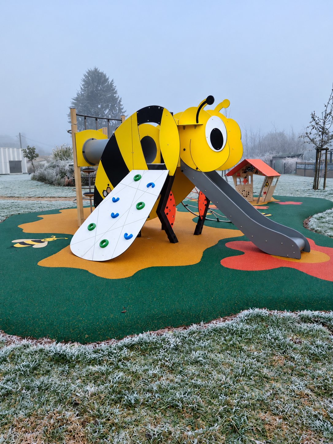 Une structure de jeux en forme de grosse abeille jaune et noire, avec toboggan et mur d'escalade, installée sur un sol souple vert et orange par un matin de brouillard givré.