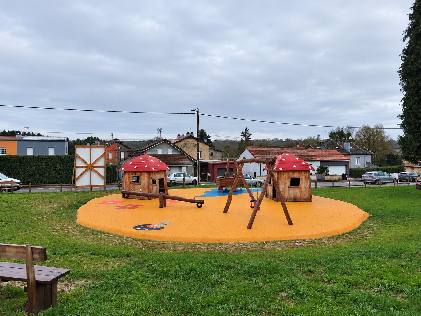 Une aire de jeux sur sol orange comprenant deux cabanes en bois à toit de champignon rouge, une balançoire et une bascule, entourée de pelouse et de maisons.
