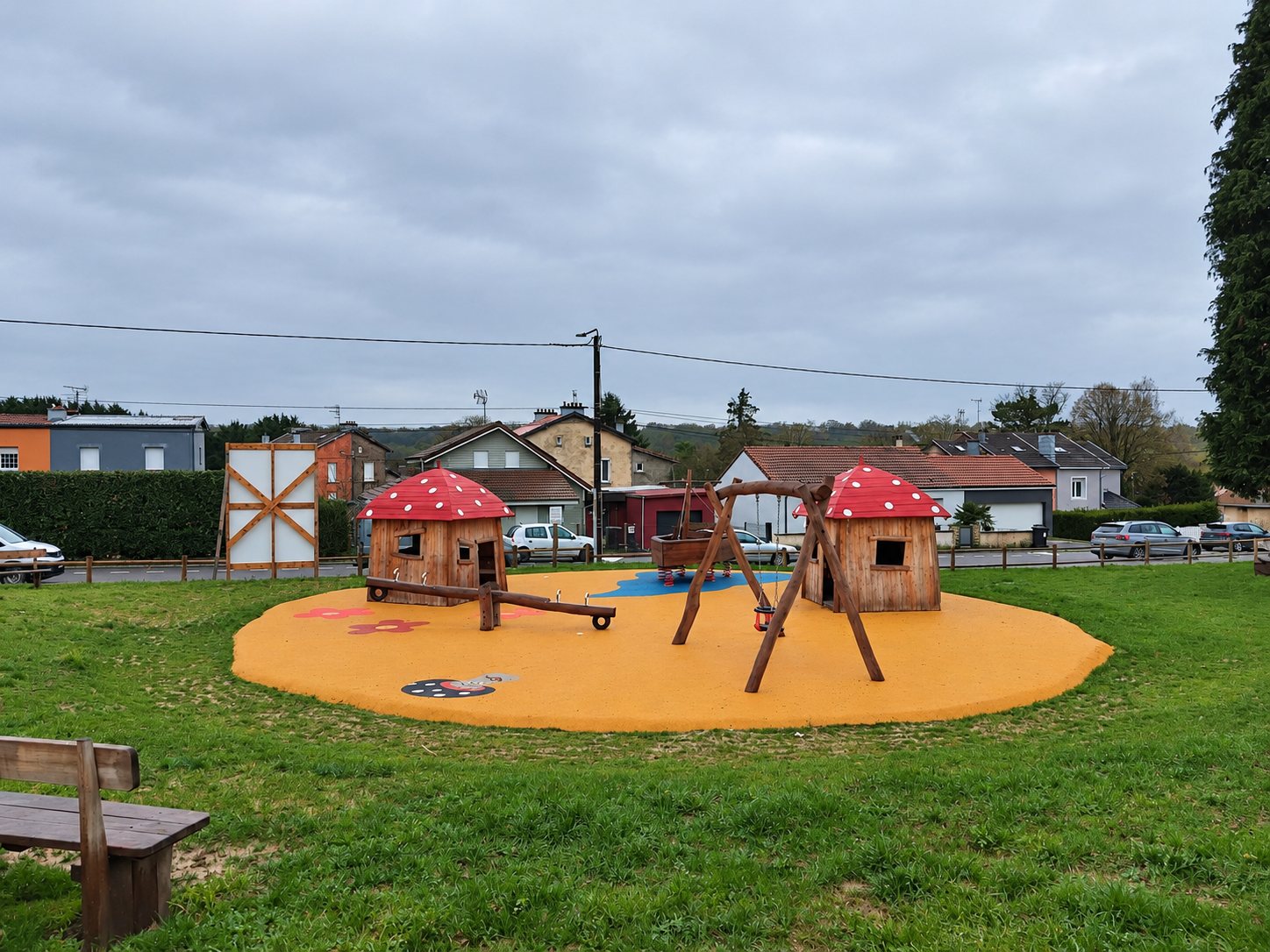 Une aire de jeux installée sur une pelouse, avec deux cabanes en bois à toit de champignon rouge, une balançoire et une bascule sur un sol souple orange.