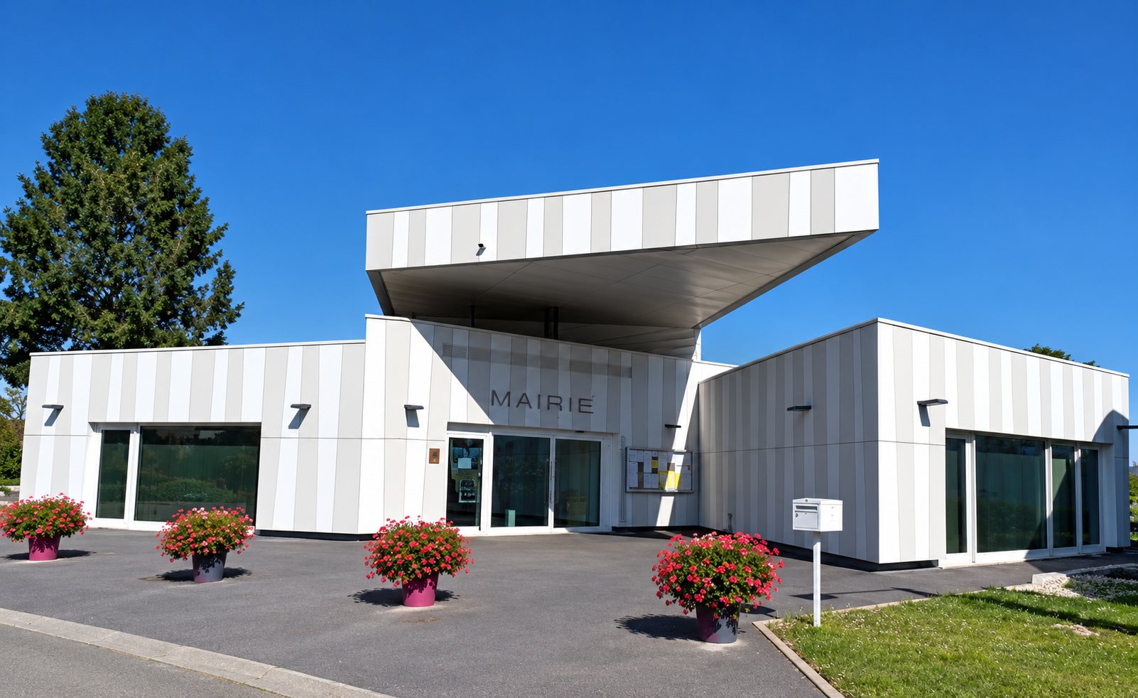 Façade d'une mairie moderne à toit plat avançant en auvent, avec des jardinières de fleurs rouges devant l'entrée sous un ciel bleu.
