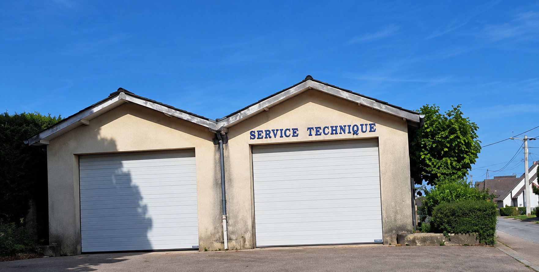 Un bâtiment aux murs beige clair avec deux grandes portes de garage blanches et l'inscription « Service Technique » au-dessus de l'une d'elles.
