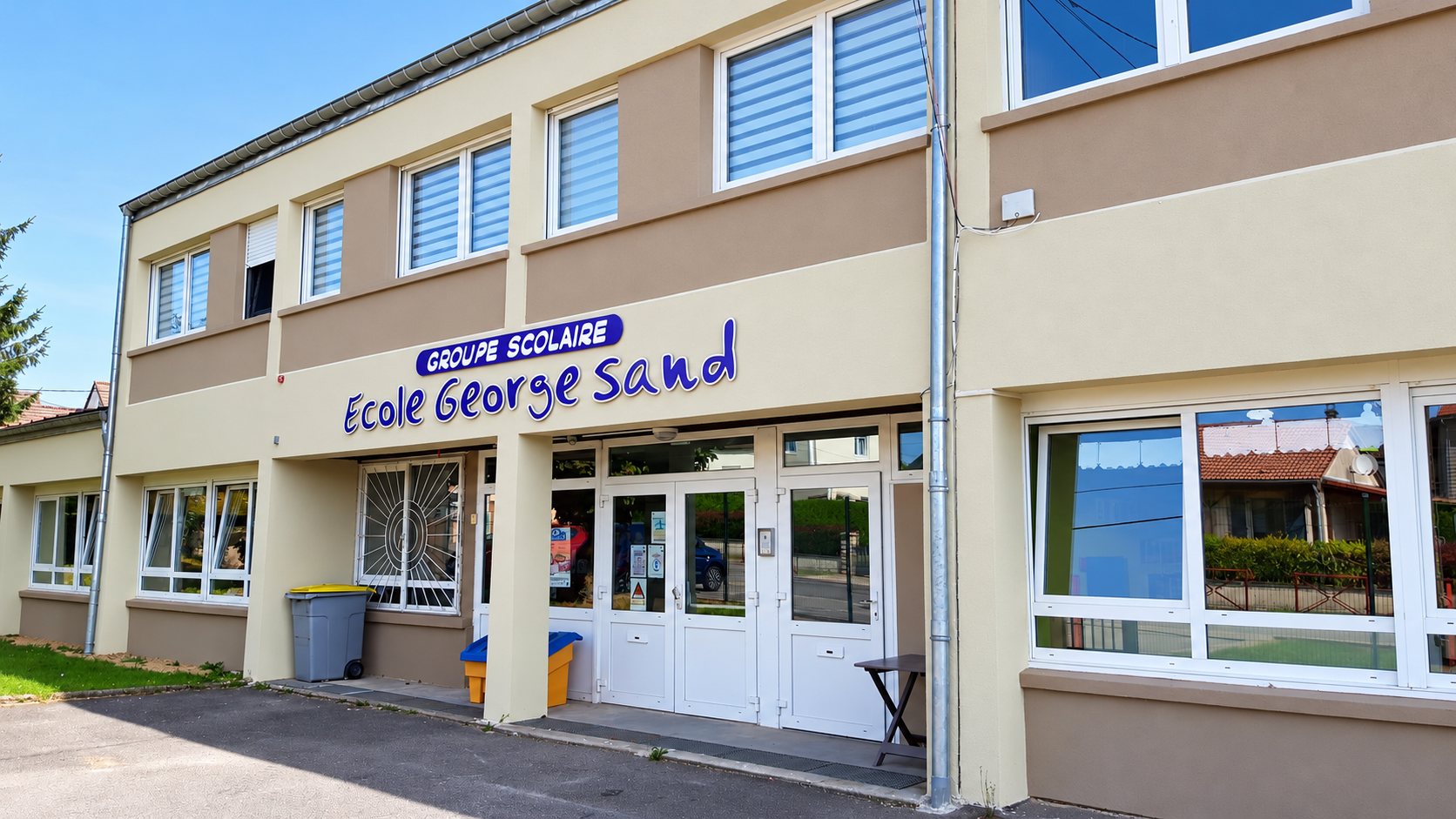 Façade beige et taupe du groupe scolaire École George Sand, avec l'entrée vitrée, des fenêtres à stores et une poubelle grise devant le bâtiment.