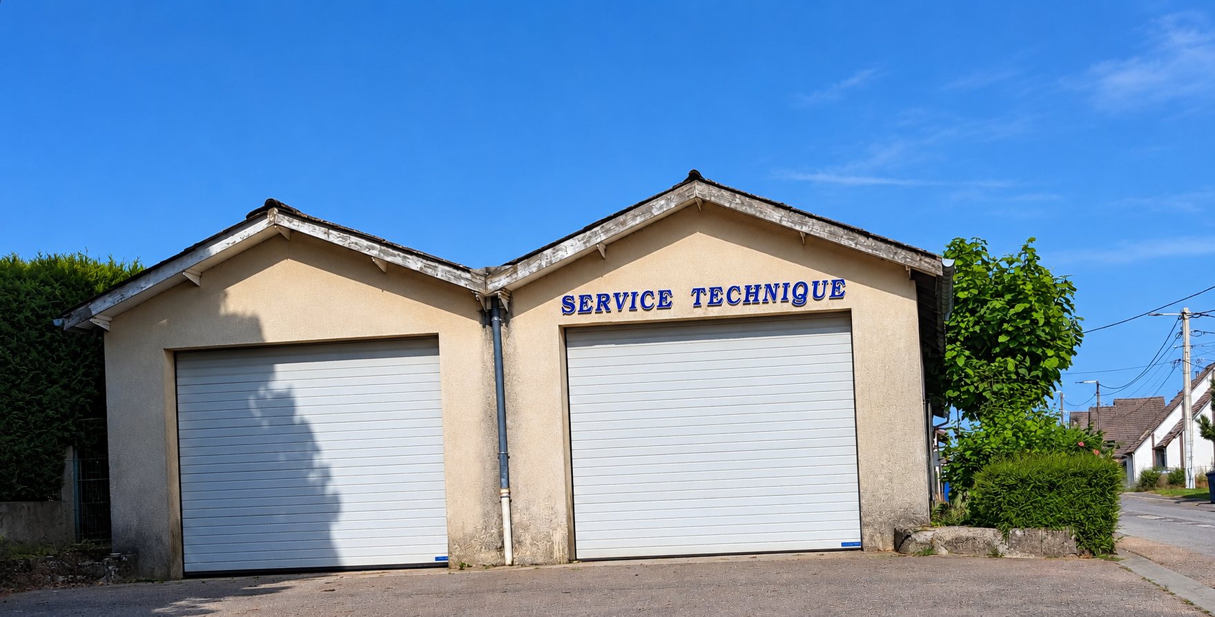 Un bâtiment de plain-pied à deux portes de garage blanches, portant l'inscription bleue « Service Technique » sous un ciel dégagé.