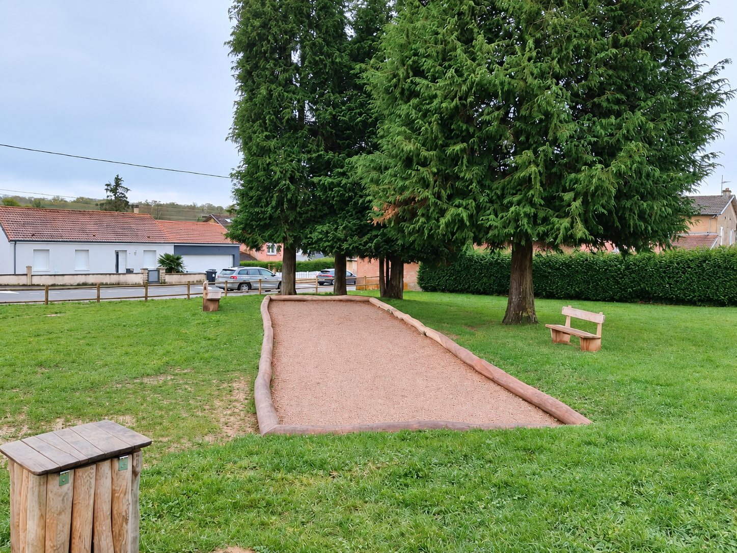 Un terrain de pétanque en gravier bordé de rondins de bois, entouré d'herbe, de bancs et de grands conifères, avec des maisons en arrière-plan.
