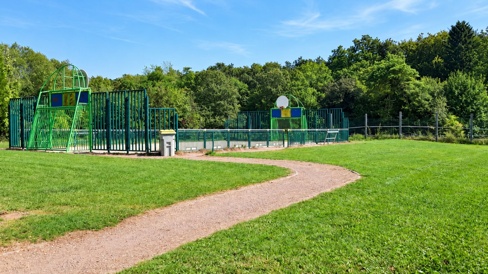 Un chemin de gravier longe une pelouse et mène à un terrain multisport entouré de barrières vertes, avec des paniers de basket et une forêt en arrière-plan.