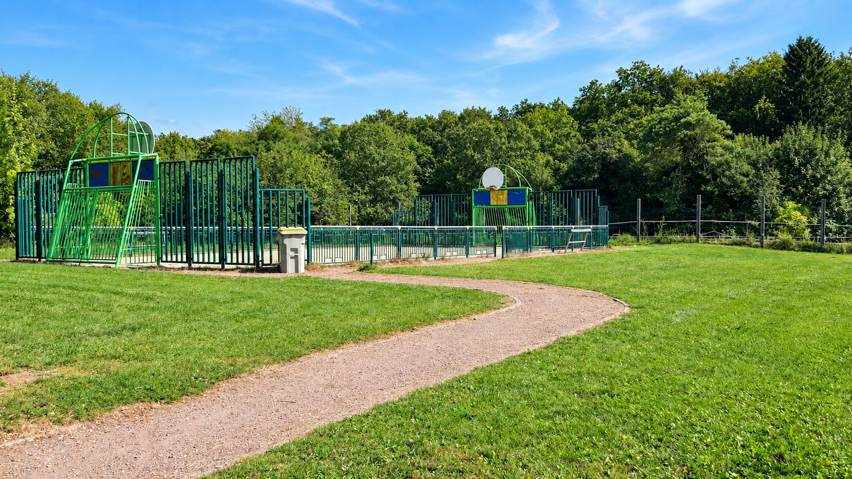 Un terrain multisport clôturé de vert avec deux panneaux de basket, accessible par un chemin de gravier sinueux traversant la pelouse, devant une forêt.
